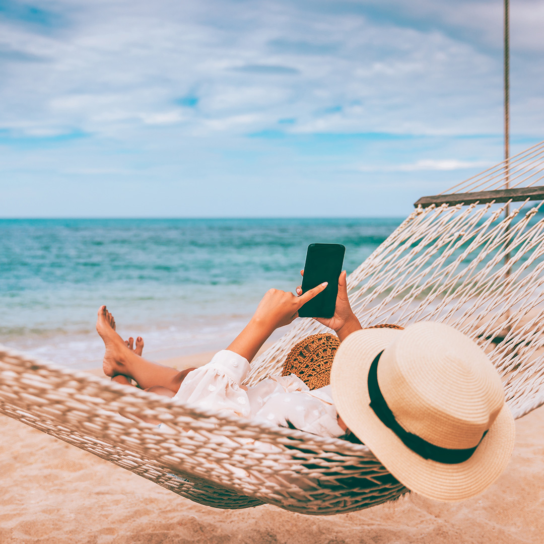 Person wearing a wide-brimmed hat lying in a hammock on the beach using a smartphone with the ocean in the background.