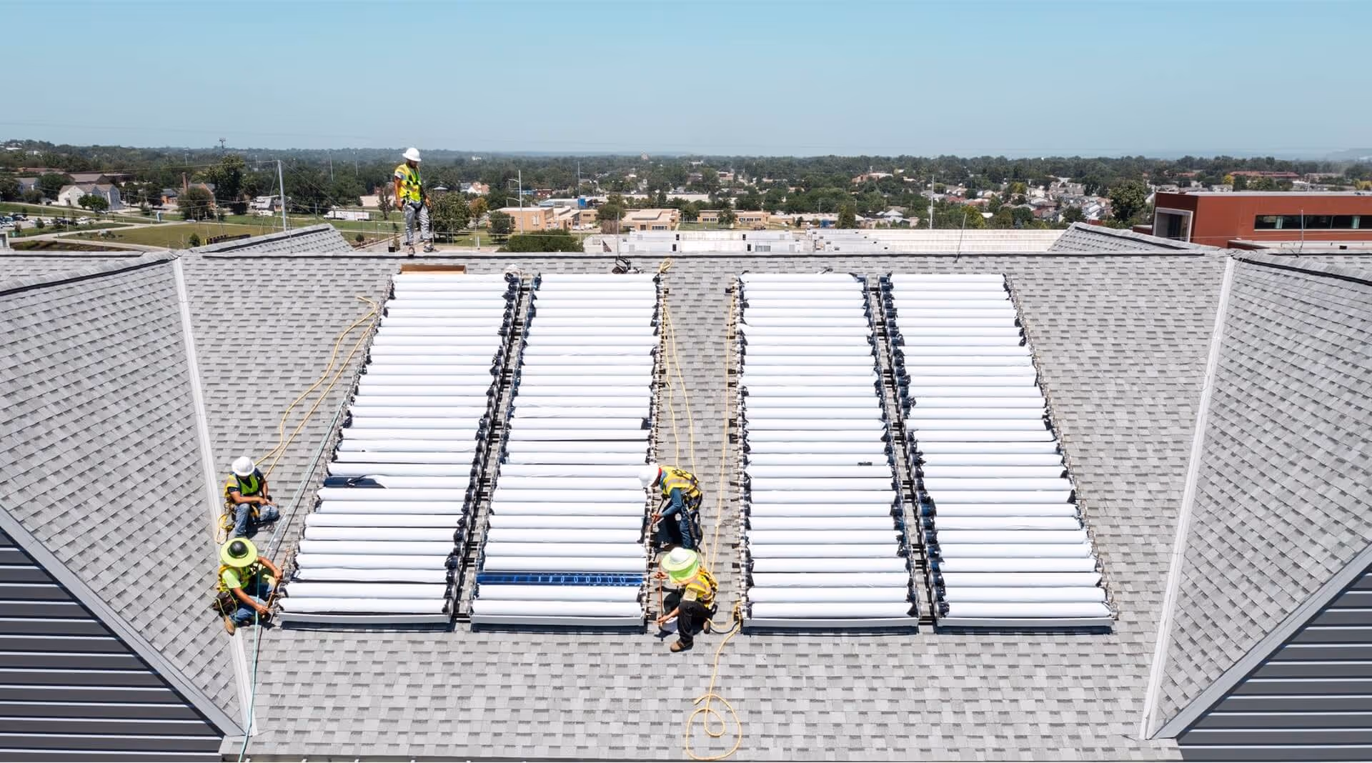 Workers installing solar thermal collectors on a gray shingled roof on a clear day.