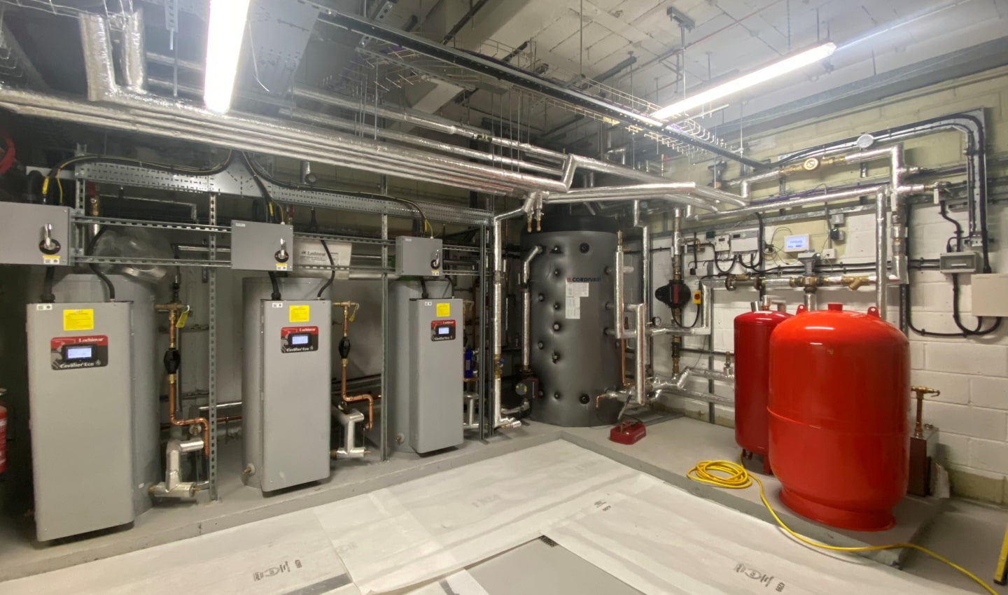 Industrial plant room with three beige heating units, a large black cylindrical tank, and two red pressure vessels surrounded by silver piping.