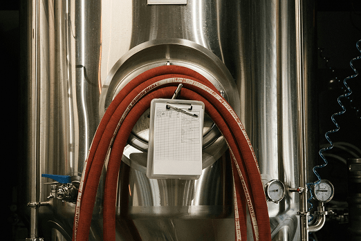 Stainless steel brewing equipment with red hoses and a clipboard attached to the front.