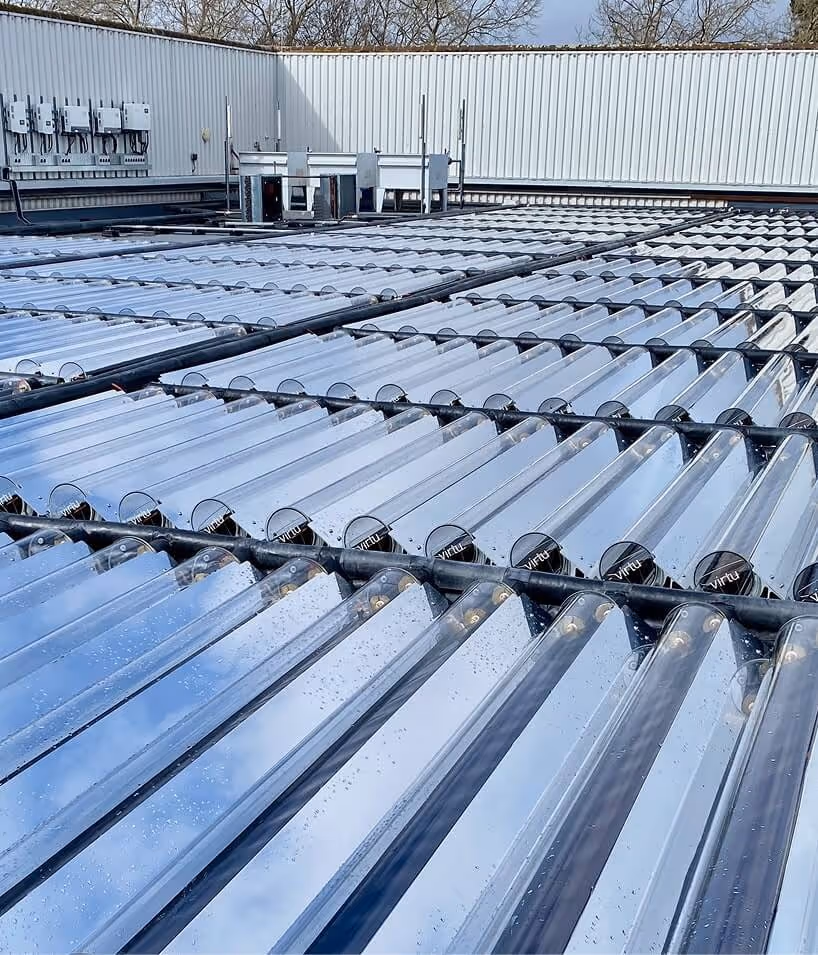 Rows of reflective solar vacuum tubes installed on a flat rooftop with electrical boxes on a surrounding wall.