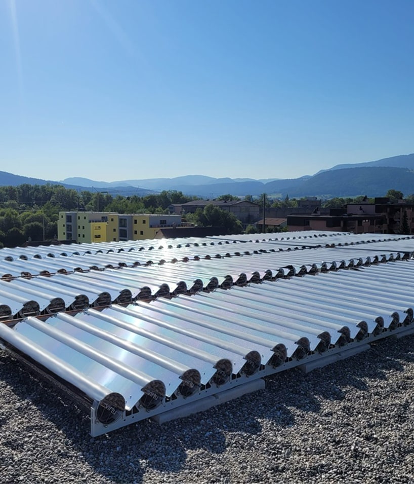 Rows of solar thermal collector tubes installed on a gravel rooftop with mountains and buildings in the background under a clear blue sky.
