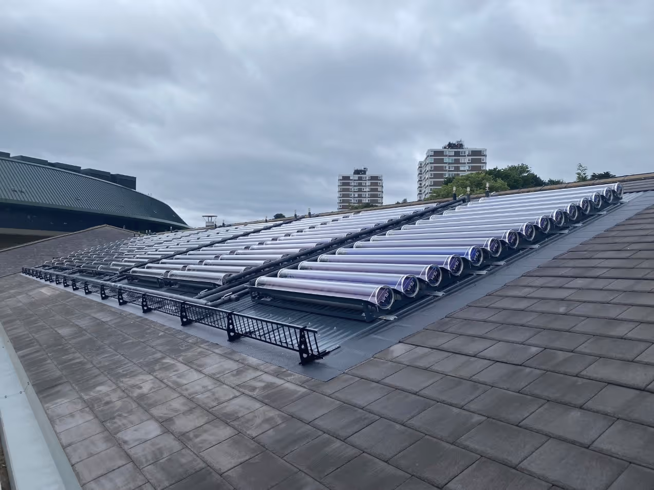 Conjunto de tubos solares al vacío instalados en un tejado inclinado de tejas grises bajo un cielo nublado, con edificios de apartamentos al fondo.