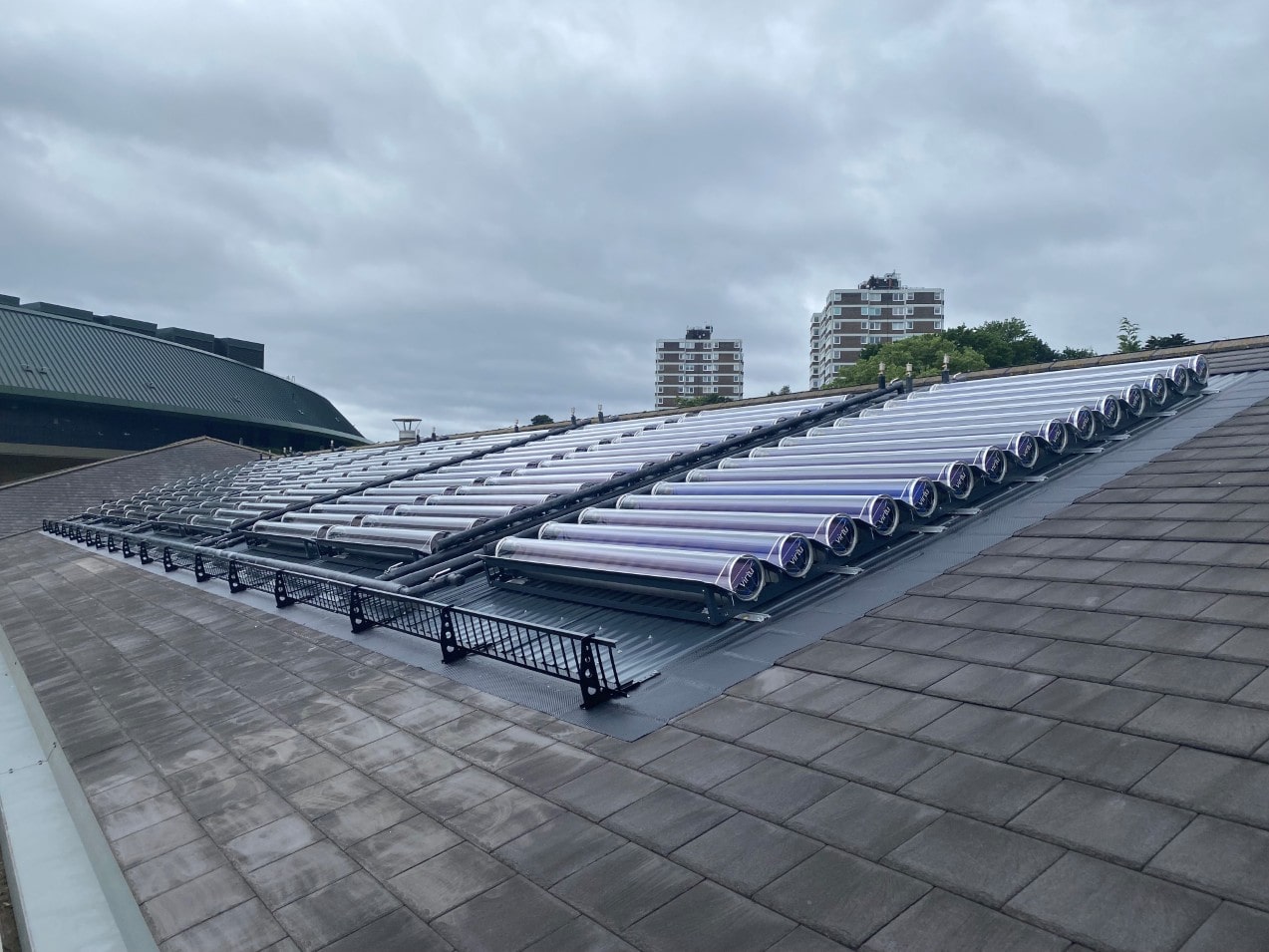 Array of solar vacuum tubes installed on a sloped grey tiled roof under a cloudy sky with apartment buildings in the background.
