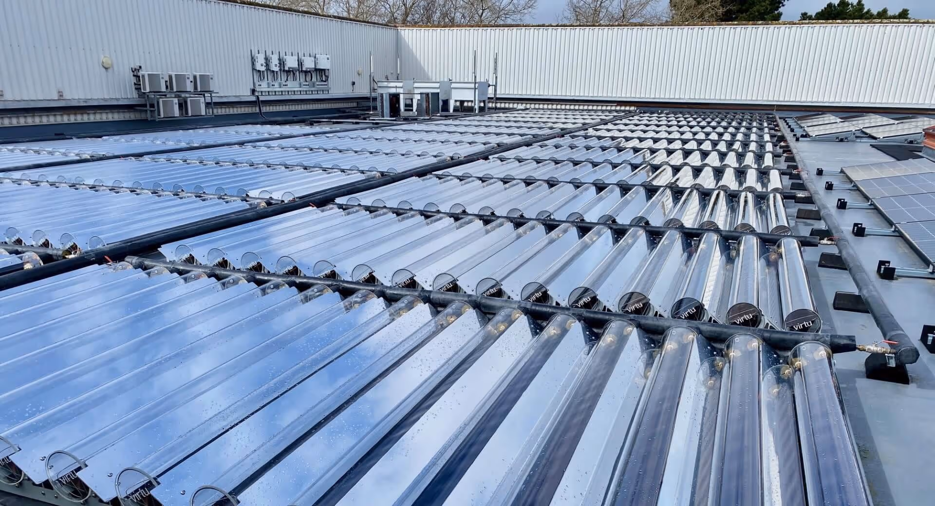 Rows of vacuum tube solar collectors installed on a flat rooftop with nearby solar panels and HVAC units.