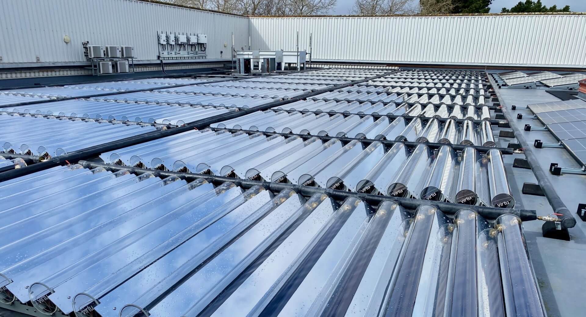 Rows of vacuum tube solar collectors installed on a flat rooftop with nearby solar panels and HVAC units.