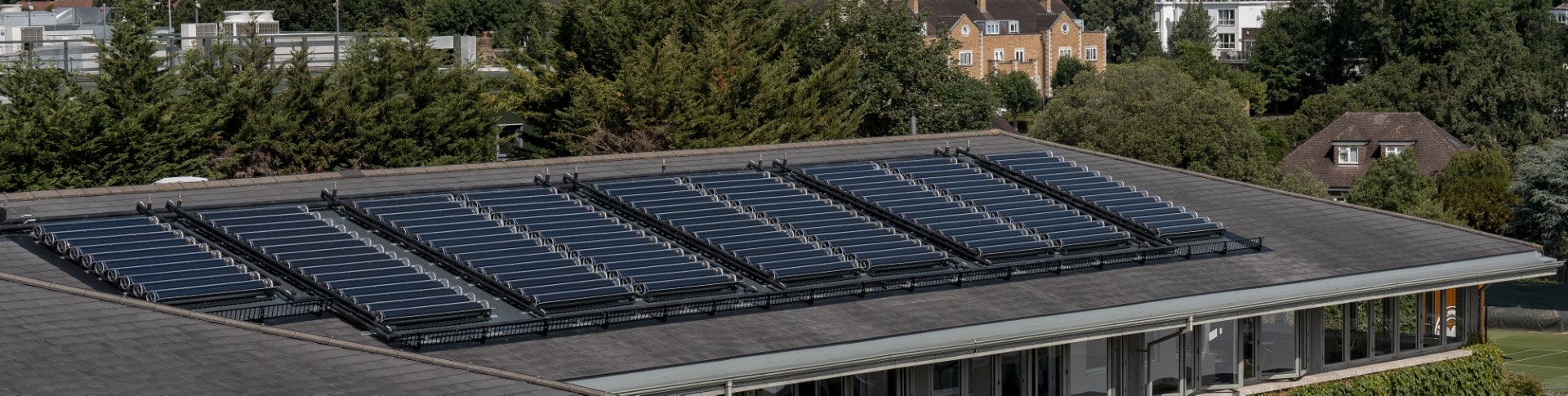 Flat rooftop with multiple rows of solar thermal panels surrounded by trees and residential buildings.
