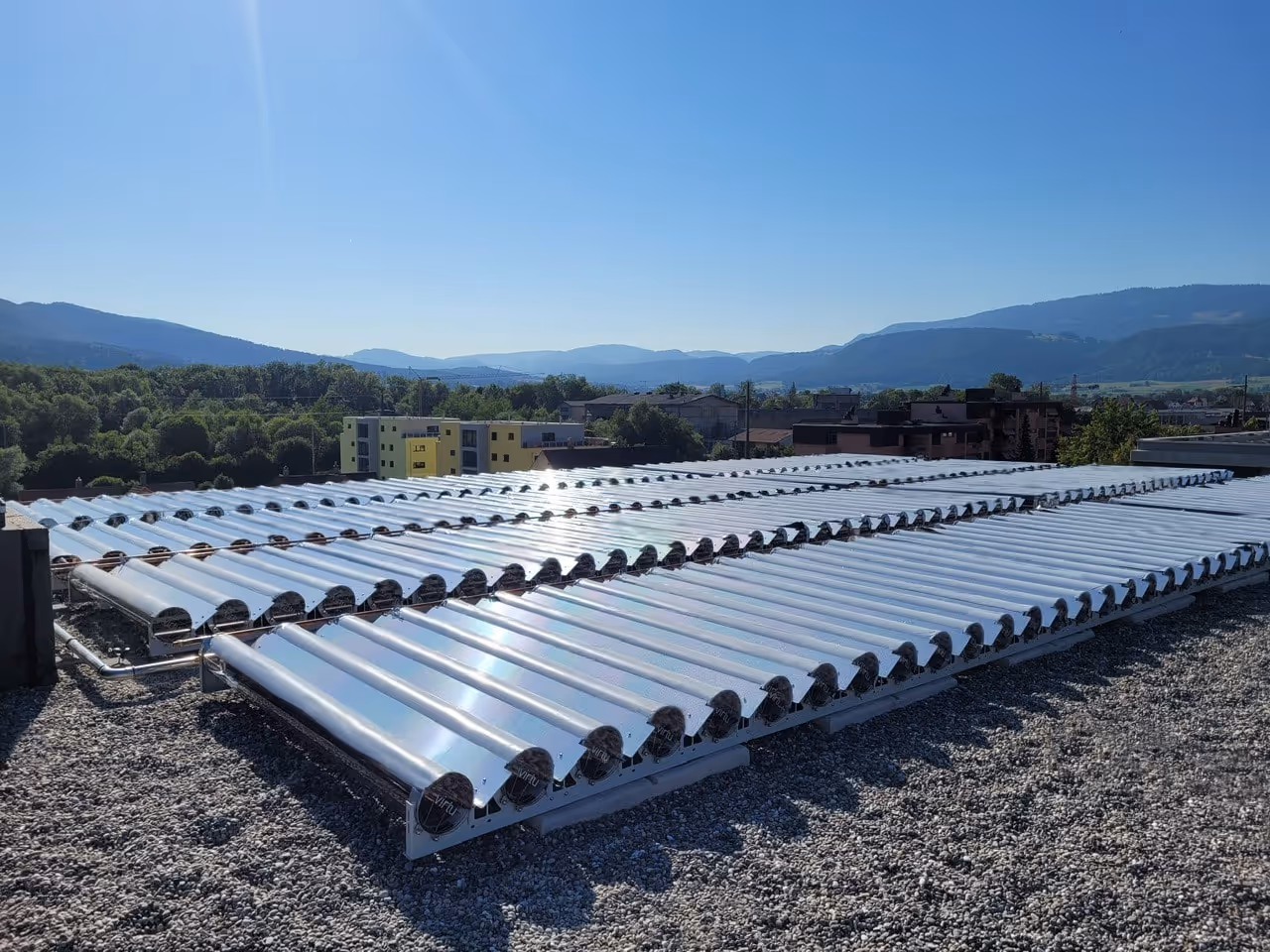 Rows of solar thermal vacuum tube collectors installed on a gravel rooftop with mountains and buildings in the distance under a clear blue sky.