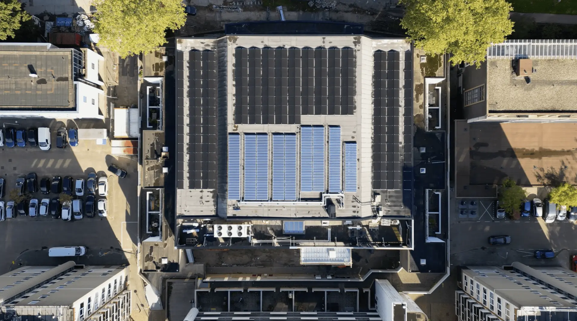 Vista aérea do telhado de um grande edifício com painéis solares e carros estacionados nas ruas circundantes.