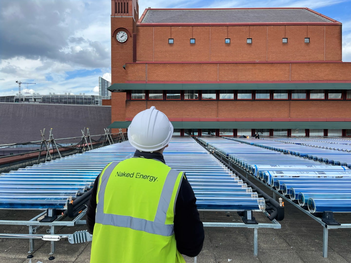 Person wearing a white hard hat and yellow vest labeled Naked Energy inspecting rows of blue solar tubes on a rooftop with a brick building and clock tower in the background.