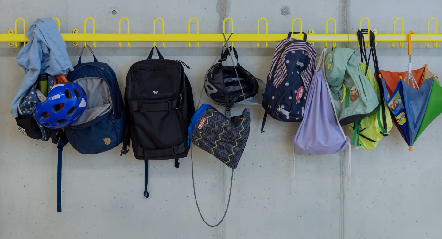 Gelbe Wandhakenleiste mit mehreren Rucksäcken, einem blauen Fahrradhelm, einer Fahrradhelm-Hülle und einem bunten Regenschirm, die an einer grauen Wand hängen.