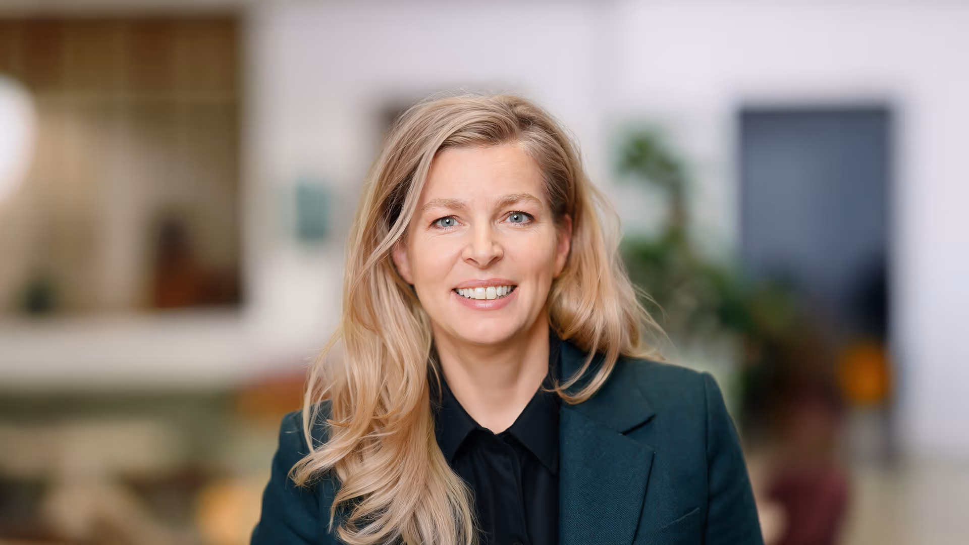 Lächelnde blonde Frau mit lockerem Haar trägt einen dunkelgrünen Blazer und schwarzes Hemd vor unscharfem Hintergrund.