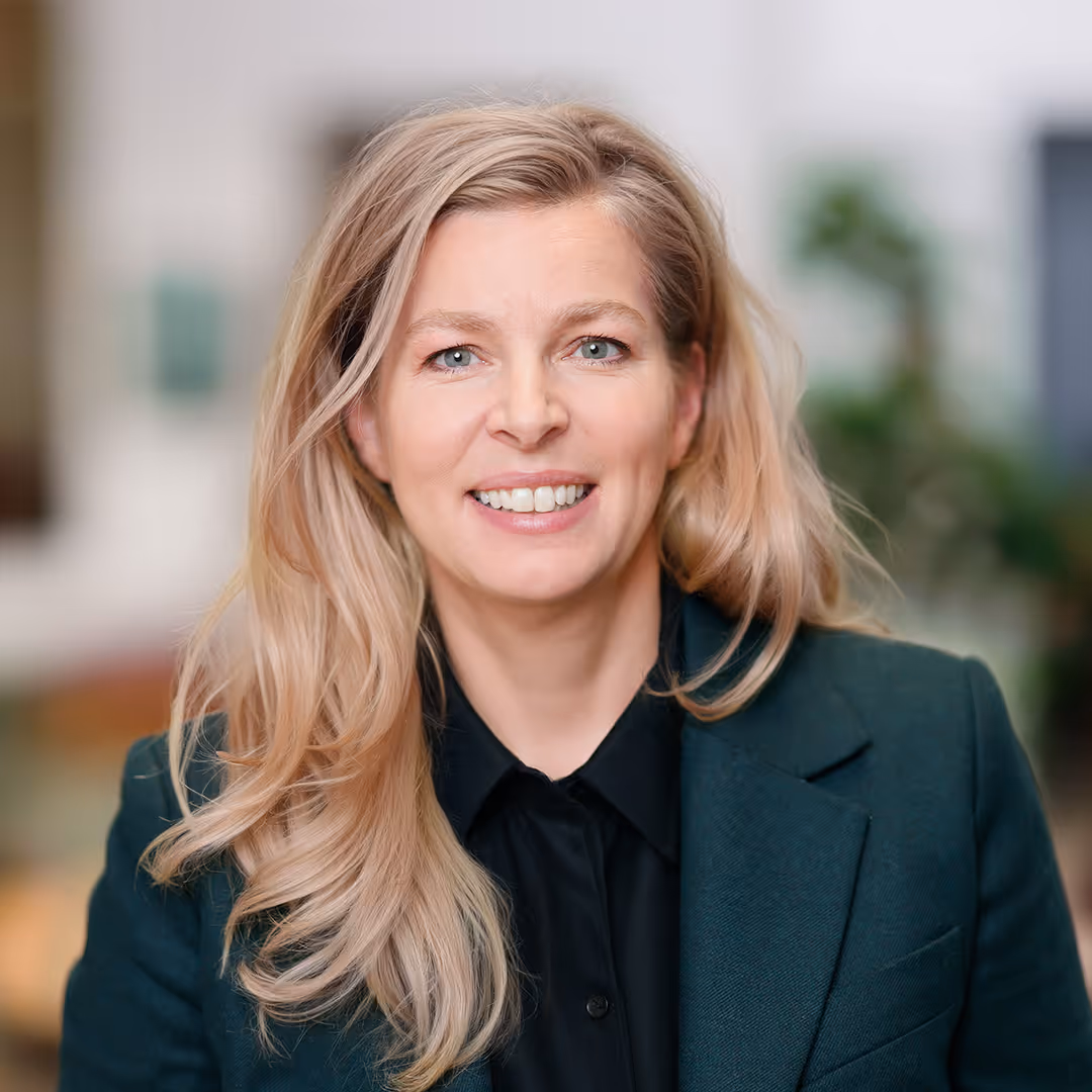 Porträt einer lächelnden Frau mit langen blonden Haaren in dunklem Blazer und schwarzem Hemd vor unscharfem Hintergrund.