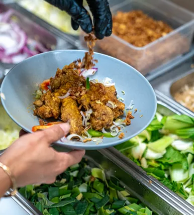 Un plat bleu rempli de morceaux frits, d'oignons rouges tranchés et de légumes frais, avec une main gantée ajoutant un ingrédient croustillant.