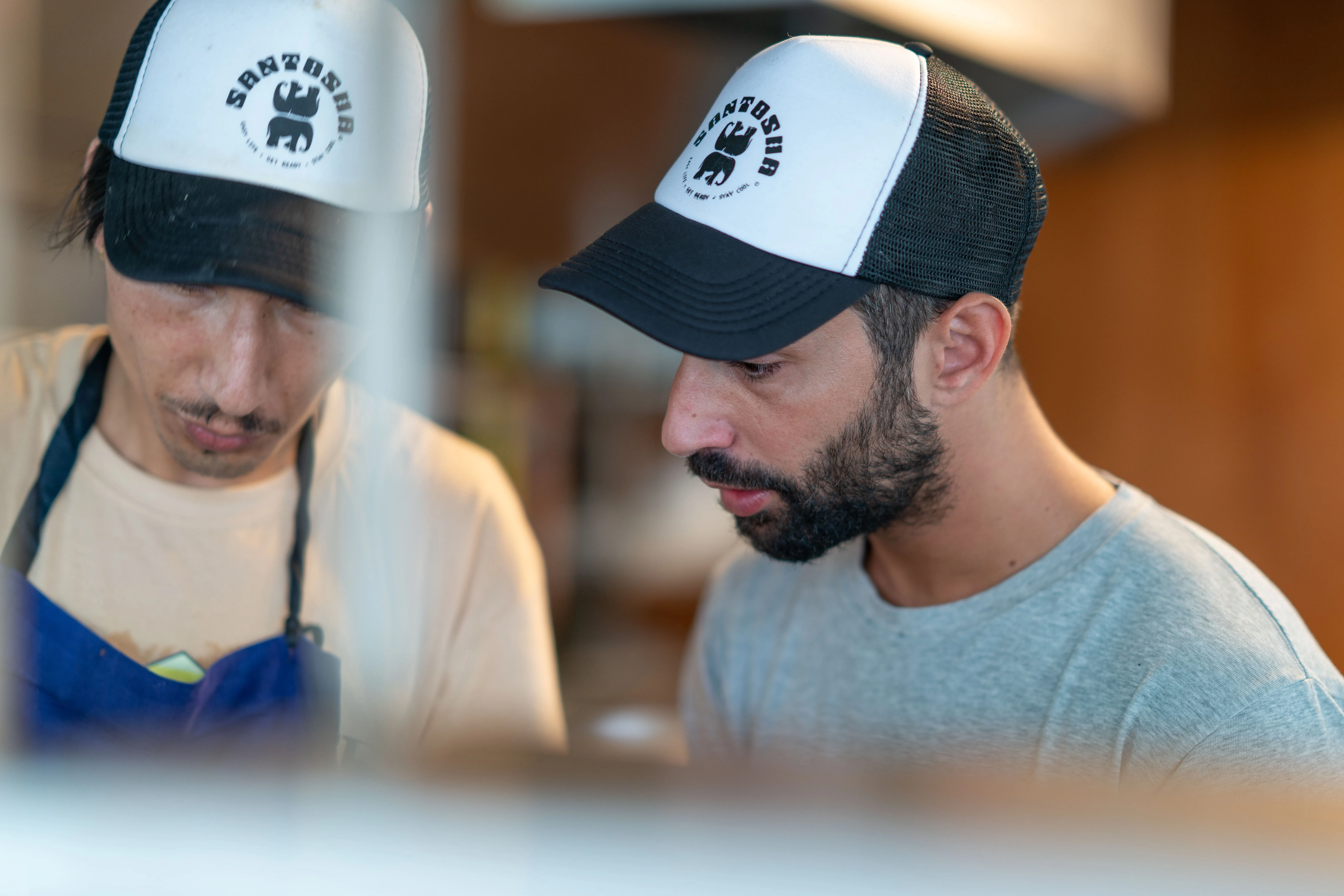 Deux hommes portant une casquette blanche et noire avec le logo Santosha, concentrés sur une tâche.