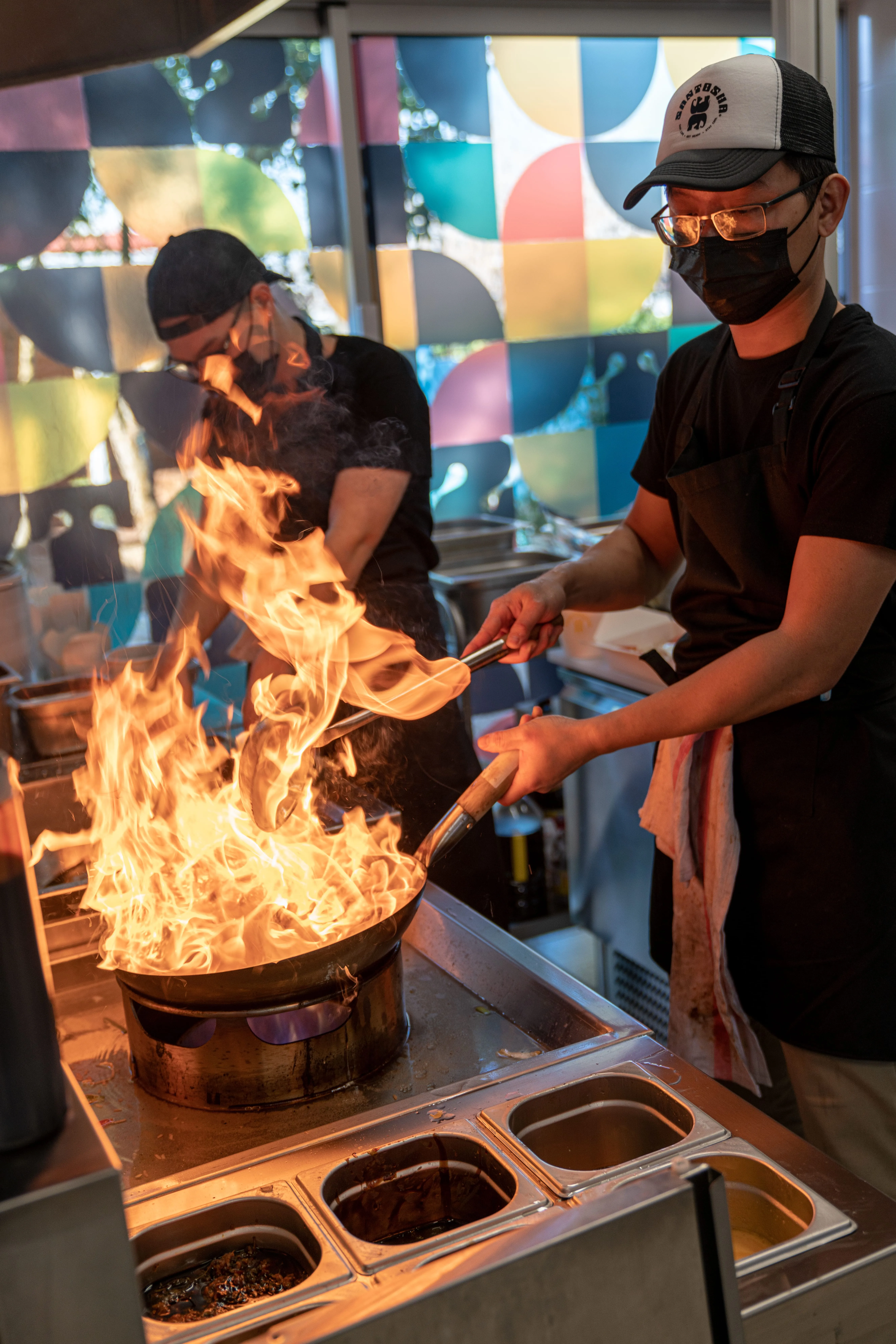 Deux cuisiniers masqués préparant des plats à feu vif dans une cuisine professionnelle avec un grand feu s'échappant d'une poêle.