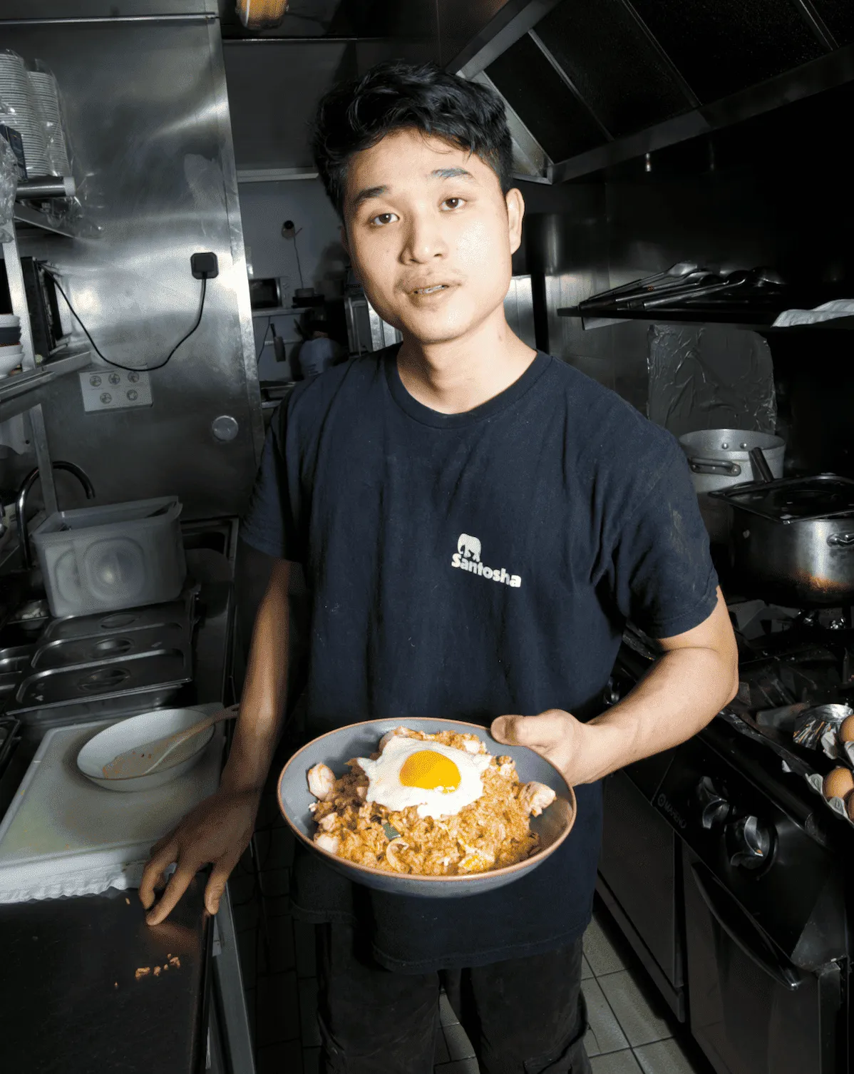 Jeune homme dans une cuisine professionnelle tenant un bol de riz sauté garni d'un œuf au plat.