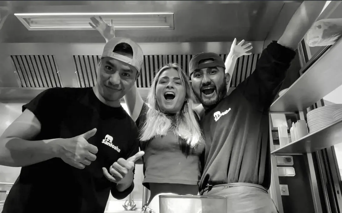 Trois jeunes adultes souriants dans une cuisine, deux hommes portant des casquettes et une femme au centre avec les bras levés.