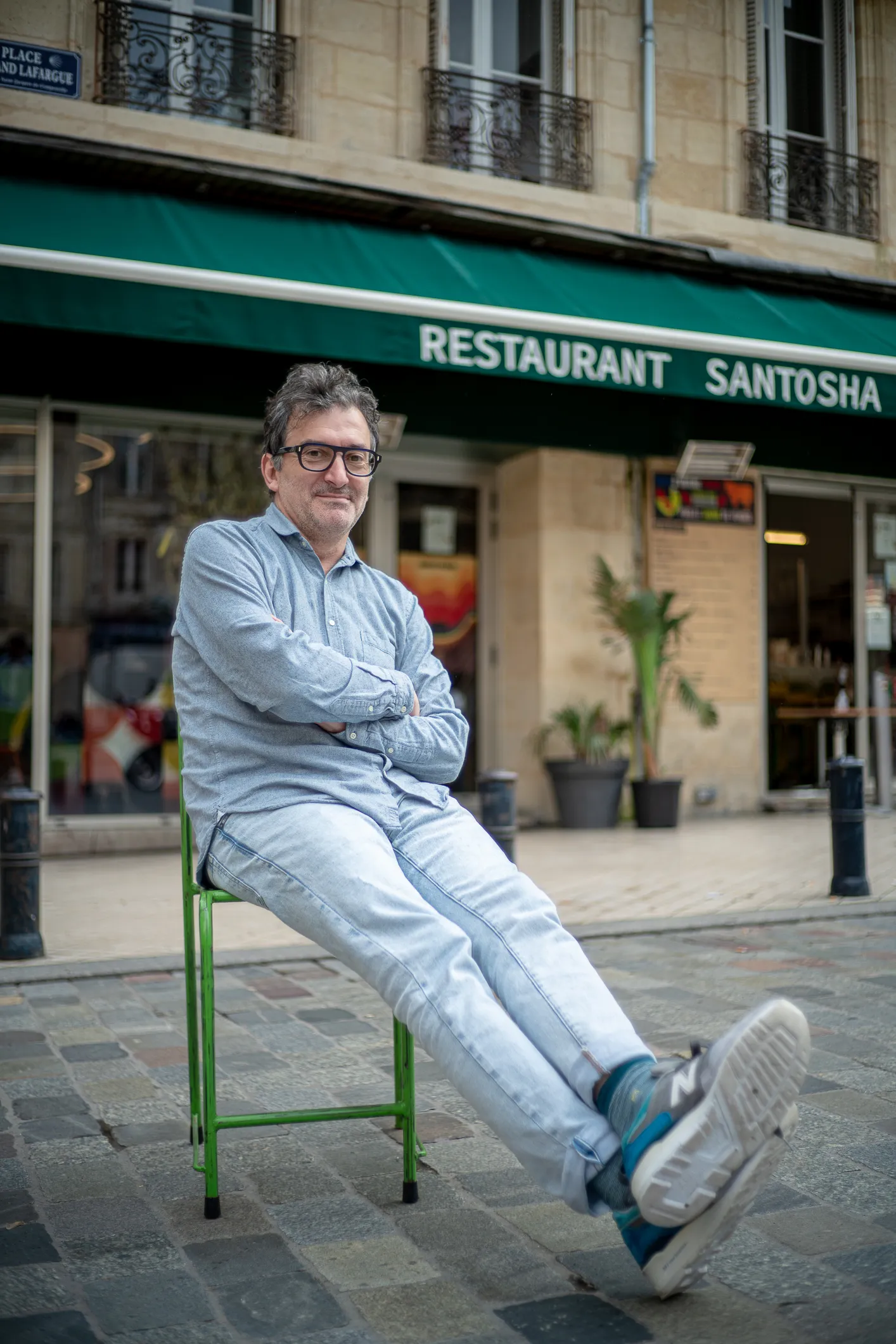 Emmanuel Meuret - fondateur de santosha - assis sur une chaise verte sur une rue pavée devant le restaurant Santosha avec un store vert.