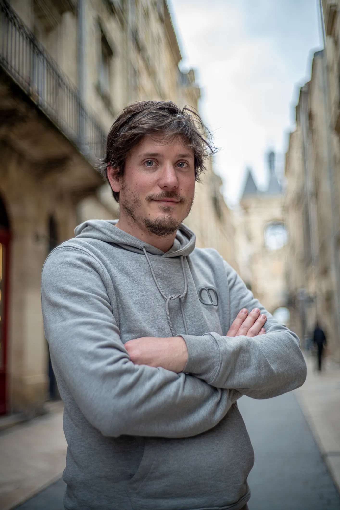 Benoit Germaneau - associe Santosha - Un homme aux cheveux bruns et à la barbe légère portant un sweat à capuche gris, bras croisés, debout dans une rue pavée entre des bâtiments anciens flous.