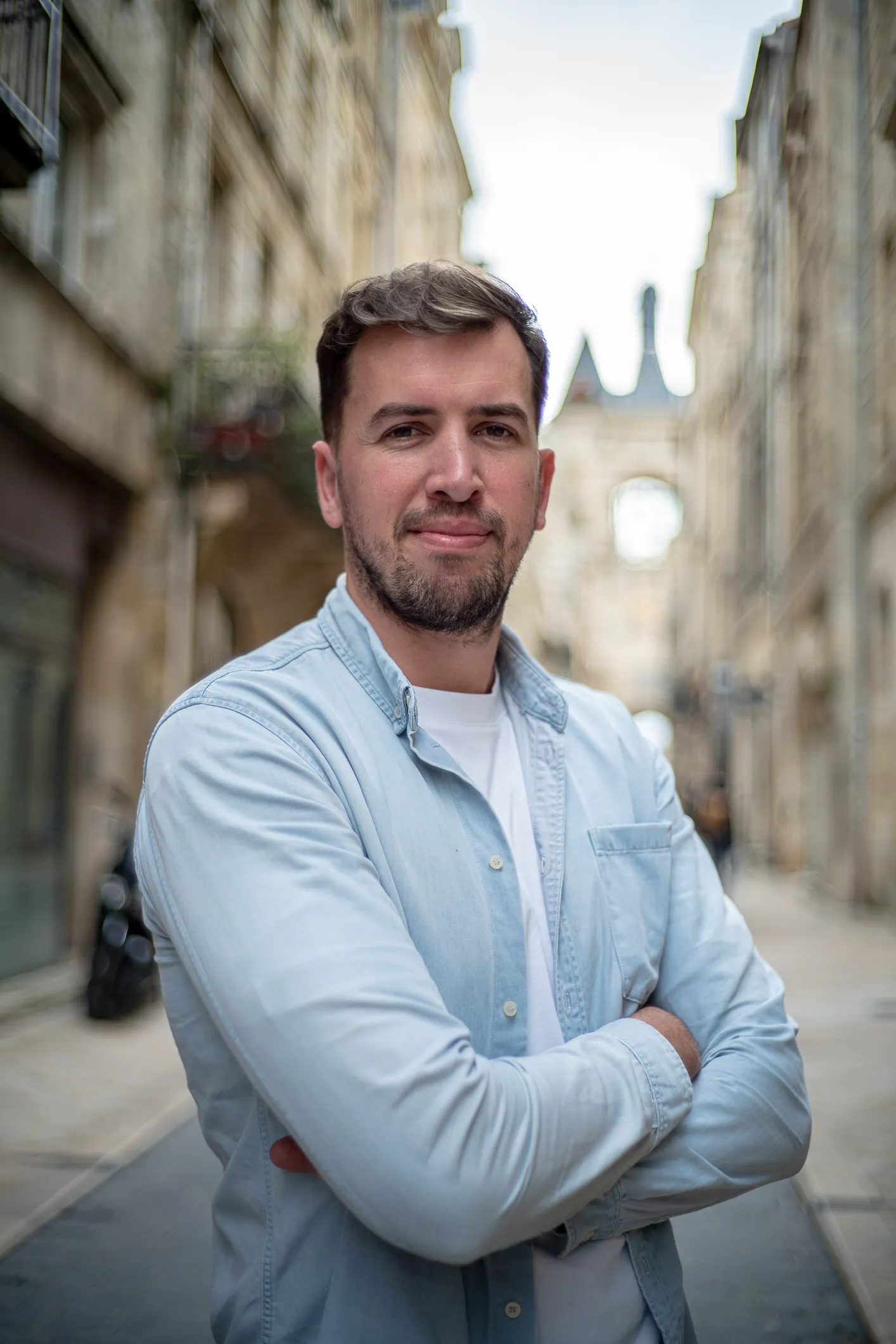 Guillaume Voidrot - associe Santosha - souriant avec chemise bleue claire et t-shirt blanc, bras croisés dans une rue urbaine floue.