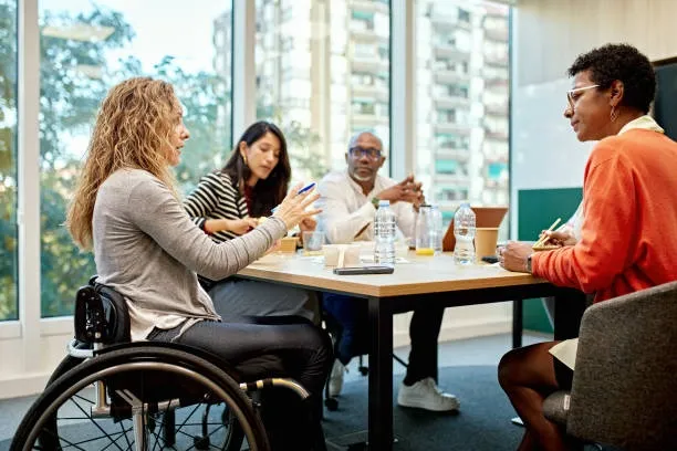Four diverse professionals engaged in a discussion around a table in a modern office, with one woman in a wheelchair.