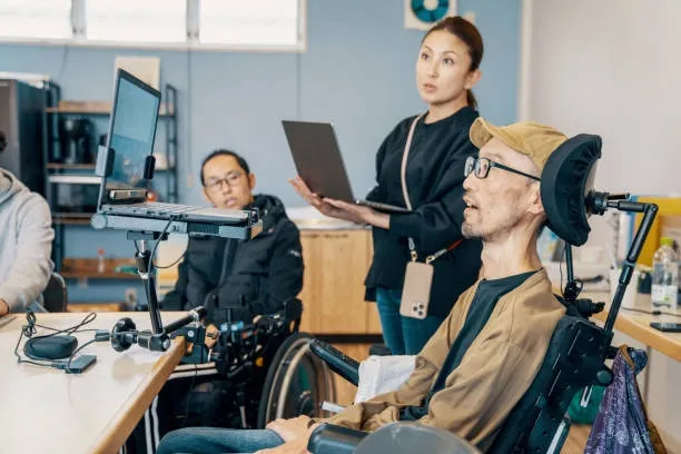 Two men in wheelchairs and a woman standing holding a laptop in a modern office setting.