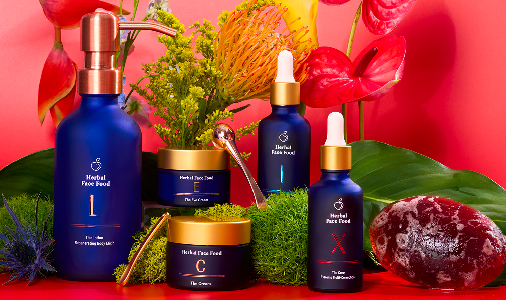 Blue bottles and jars of Herbal Face Food skincare products with gold lids, surrounded by green moss, tropical leaves, red and yellow flowers, and a textured stone on a red background.