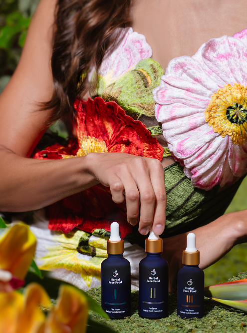 Woman in floral dress holding and displaying three dark blue bottles of herbal face food serum with dropper caps on a mossy surface.