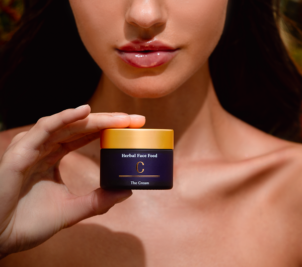 Close-up of a woman with glossy lips holding a jar of Herbal Face Food cream with a gold lid.