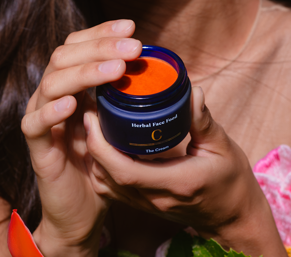 Person holding a blue jar labeled 'Herbal Face Food C The Cream' containing bright orange cream.
