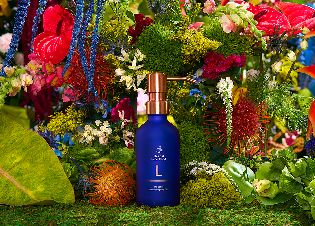 Blue bottle of Herbal Face Food lotion with gold pump surrounded by colorful flowers and green moss.
