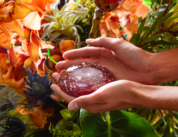 Hands holding a smooth, round, translucent purple stone with bubbles, surrounded by vibrant orange and green flowers and foliage.