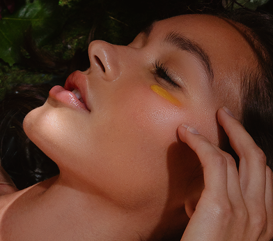 Close-up of a woman with smooth skin and a yellow paint streak under her eye, touching her face gently.