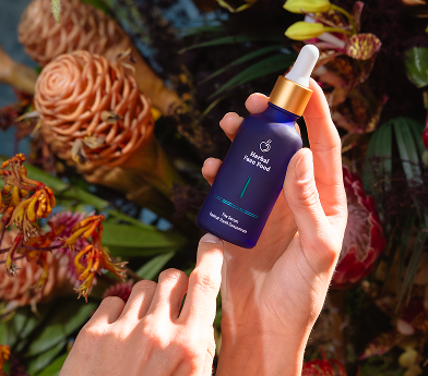 Hands holding a blue dropper bottle labeled 'Herbal Face Food' surrounded by colorful flowers and greenery.