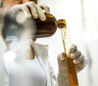 Person wearing gloves pouring amber liquid from a brown bottle into a graduated cylinder in a laboratory setting.