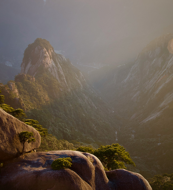 Sunlit mountain valley with rocky peaks and scattered green trees under soft hazy light.