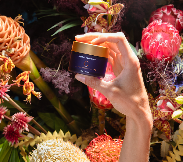 Hand holding a jar of Herbal Face Food cream surrounded by colorful flowers.