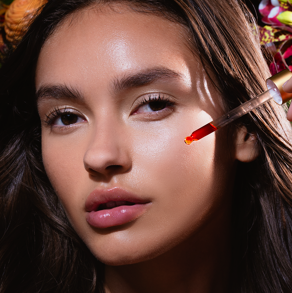 Close-up of a woman applying red facial serum with a dropper to her cheek.