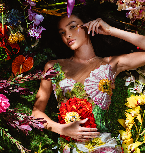 Woman lying surrounded by colorful flowers, wearing a dress with large floral designs while applying cream near her eye.