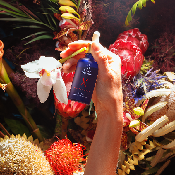 Hand holding a blue bottle of Herbal Face Fond with a dropper among colorful tropical flowers and plants.