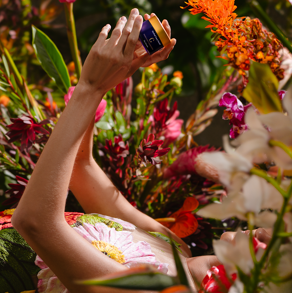 Person lying among colorful flowers holding a small jar of cream with a purple label and orange lid.