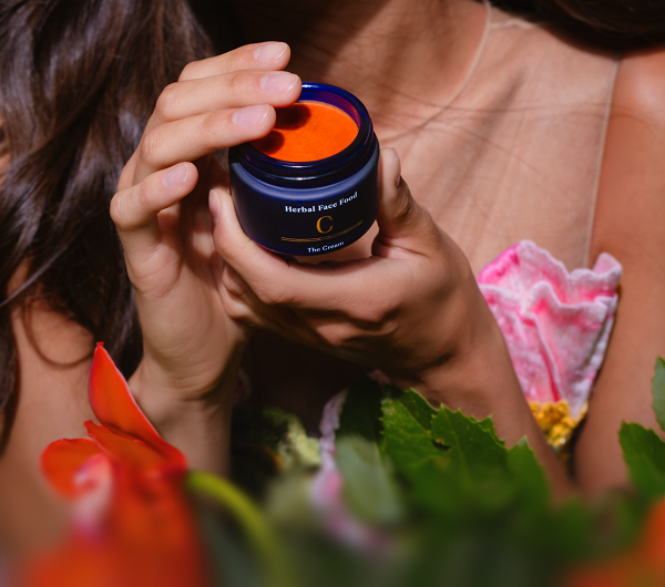 Woman holding an open jar of orange herbal face cream labeled 'Herbal Face Food C The Cream'.
