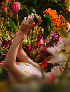 Person holding a small jar of cream surrounded by colorful flowers.