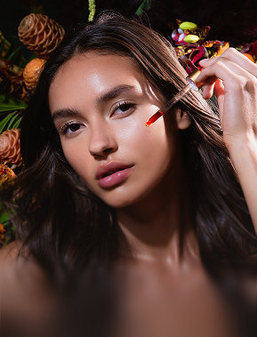 Close-up of woman with long dark hair applying red serum to her cheek using a dropper, with pine cones and colorful flowers in the background.