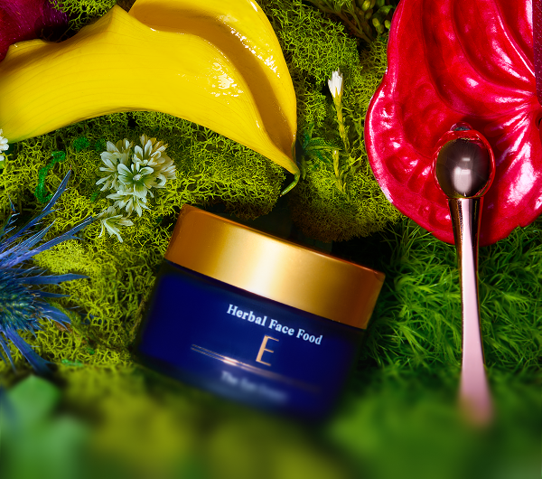 Blue jar of Herbal Face Food with a gold lid placed on green moss, surrounded by yellow and red tropical leaves and flowers with a small metallic spoon.