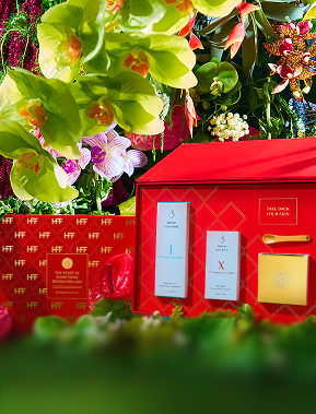 Luxury gift set featuring red boxes with gold accents and two skincare product bottles surrounded by green and pink flowers.