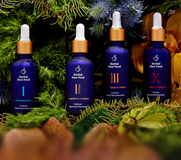 Four blue frosted glass bottles of Herbal Face Food serums with gold droppers, arranged among green and purple foliage.