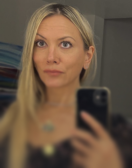 Woman with long blonde hair taking a selfie with a smartphone in front of a mirror.