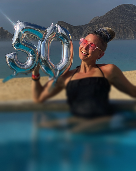 Smiling woman in a pool holding silver balloons shaped as the number 50 with a beach and rocky hills in the background.